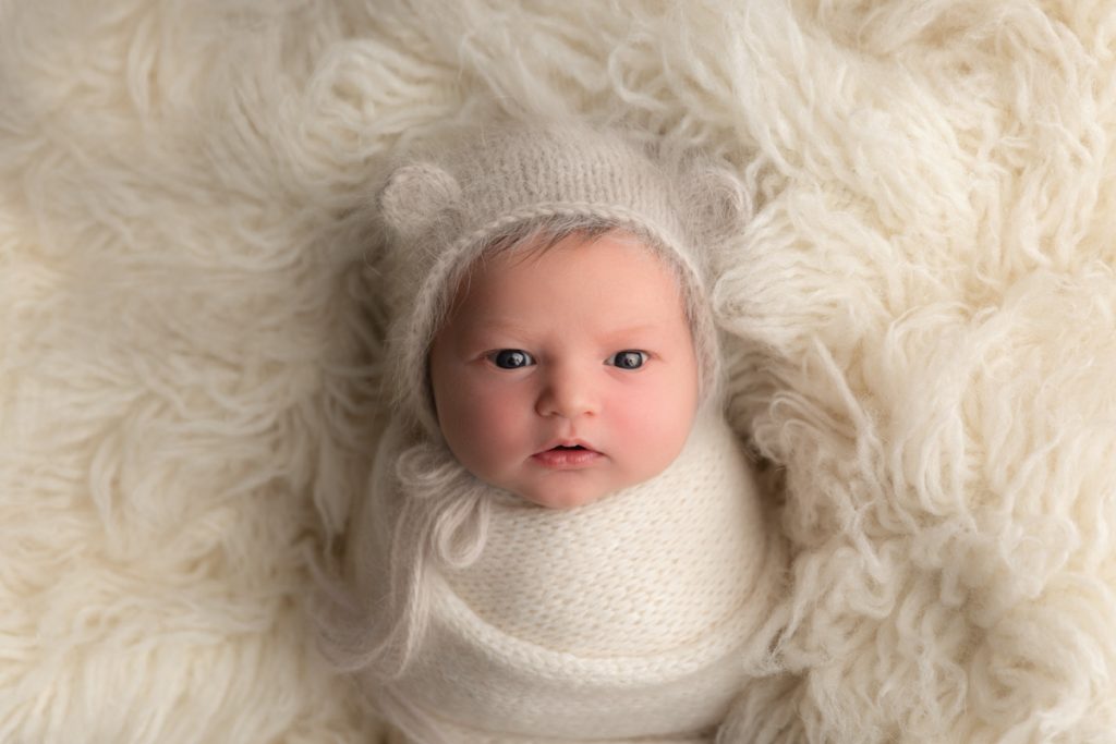 newborn baby in a teddy bear bonnet wrapped in a swaddle, resting on a fur blanket